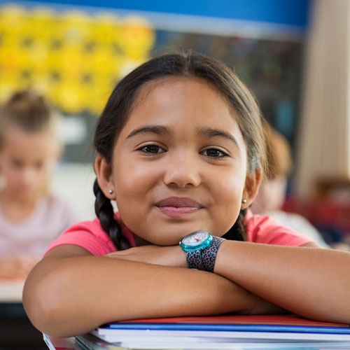 a smiling elementary school girl resting her head in her arms on a stack of books